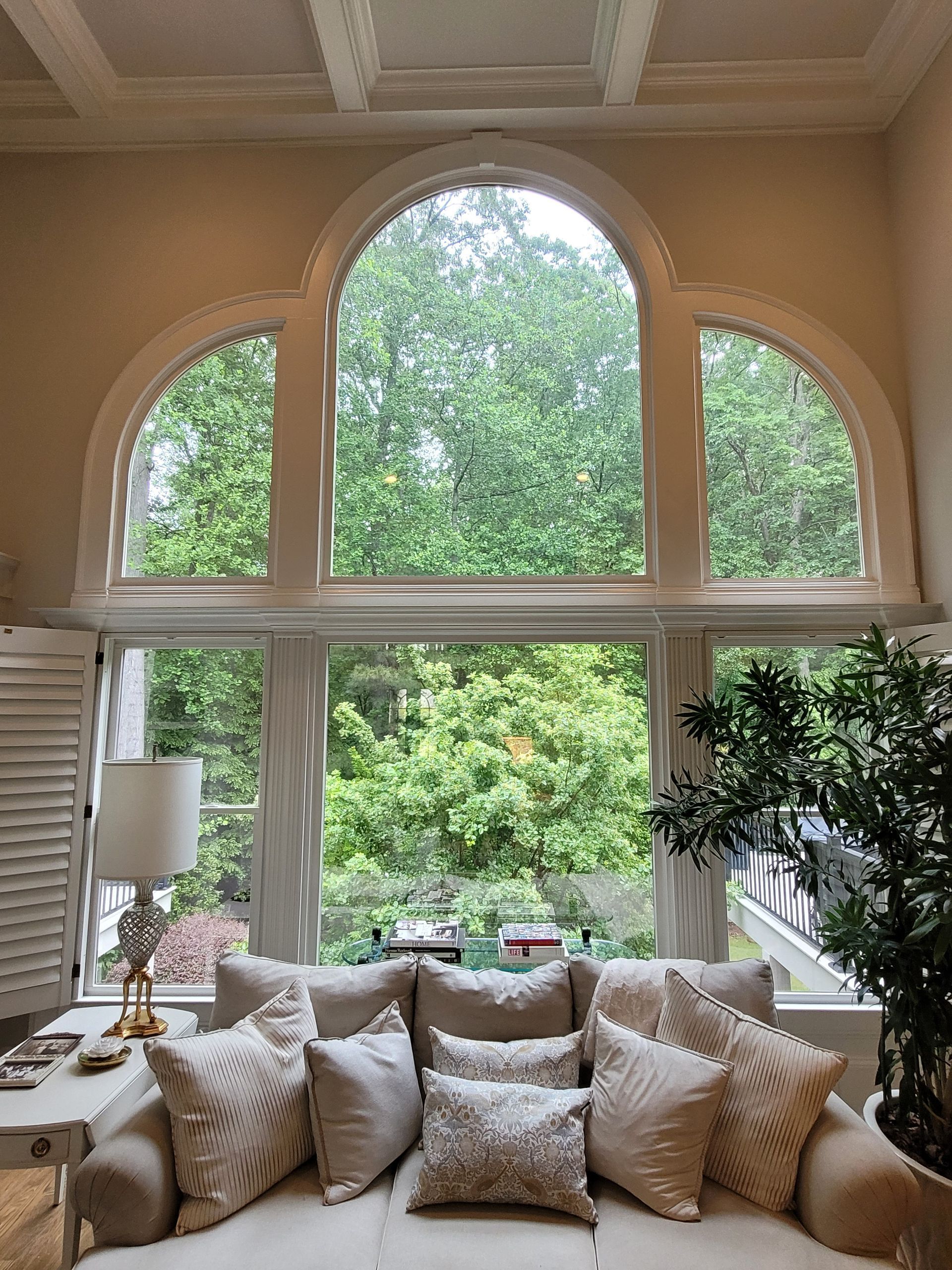 A tall, arched window frames a lush green outdoor view above a cream-colored couch with decorative pillows.