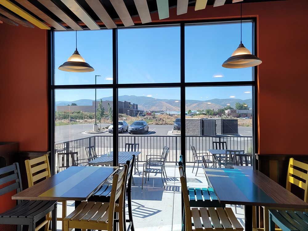 Interior view of a restaurant with large windows overlooking a patio and distant mountains. Tables and colorful chairs are visible, lit by hanging lamps.