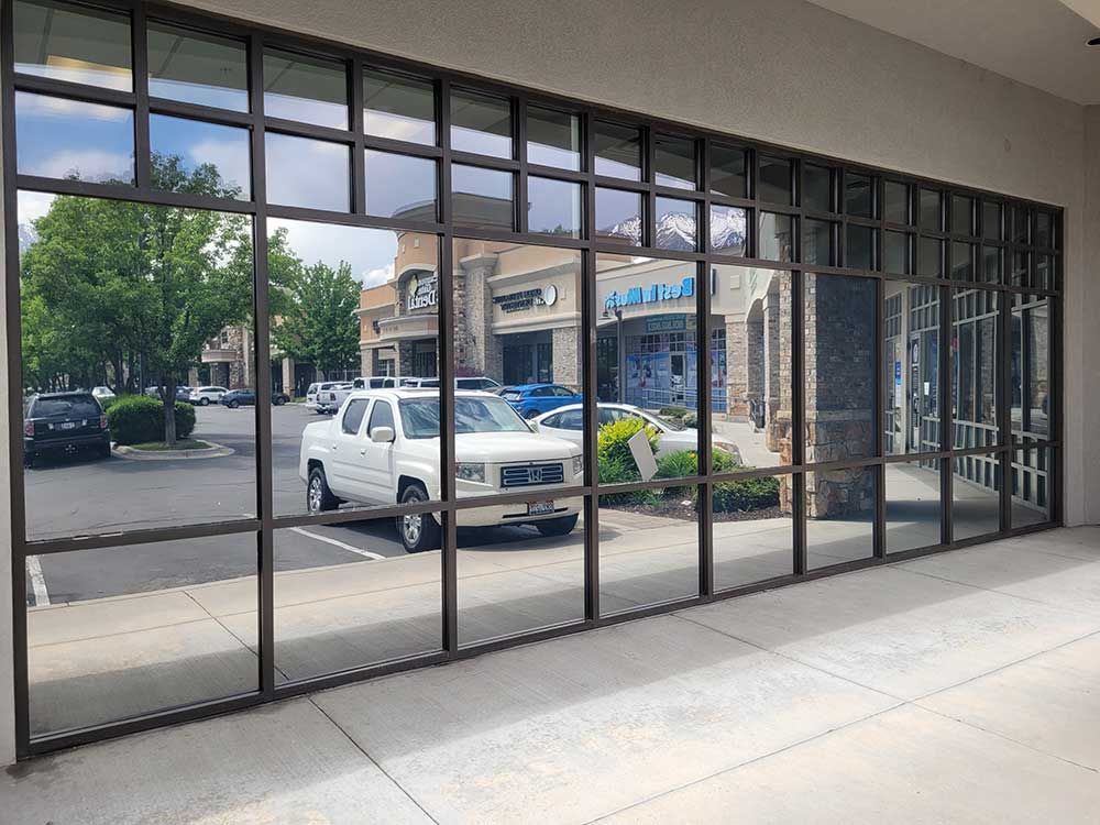 Exterior view of a building with large grid windows reflecting a parking lot with cars, trees, and other storefronts.
