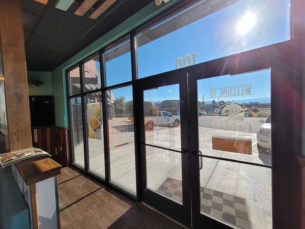 Interior view of a storefront with large windows and a bright, sunny outdoor scene visible. A wood railing and doorway are on the left.