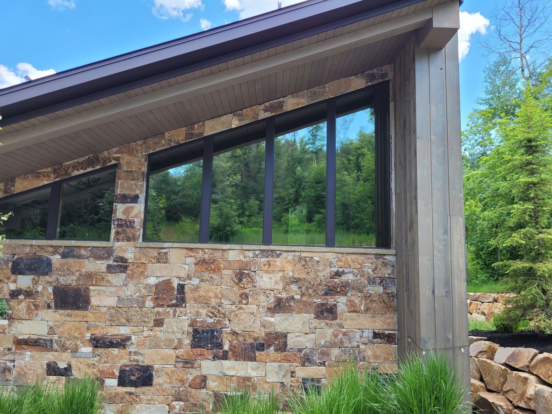 Stone-walled modern building with large, dark windows reflecting the surrounding green trees. The building has a light-colored roof and a wooden side support.