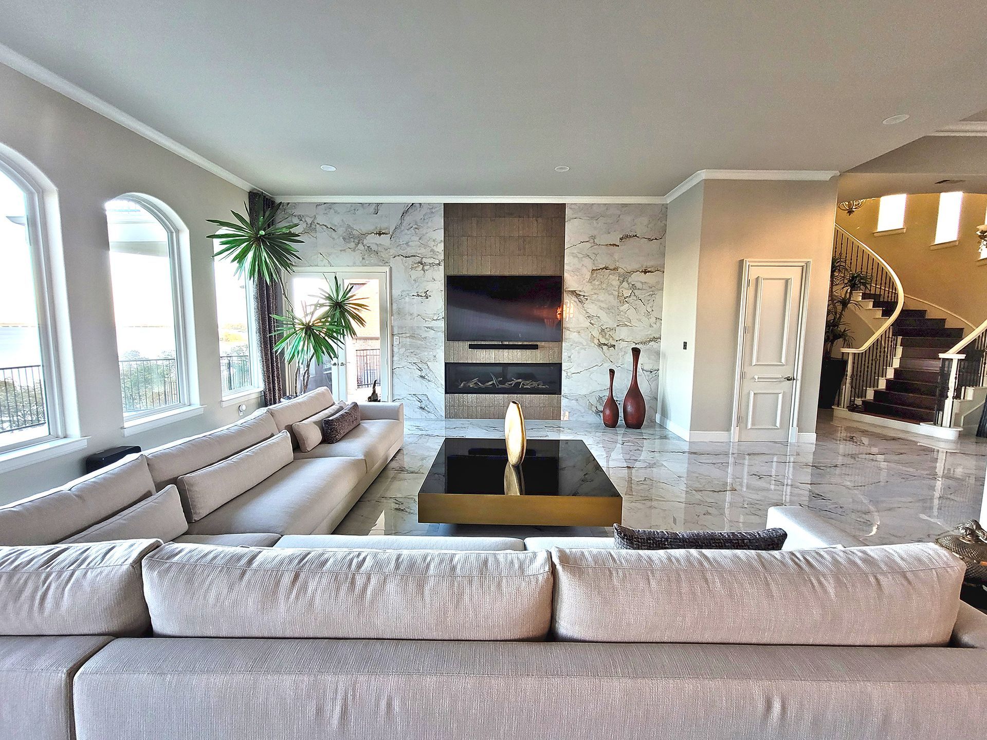 Spacious living room with large sectional sofa, fireplace, and marble floors. Sunlight streams through arched windows. - After