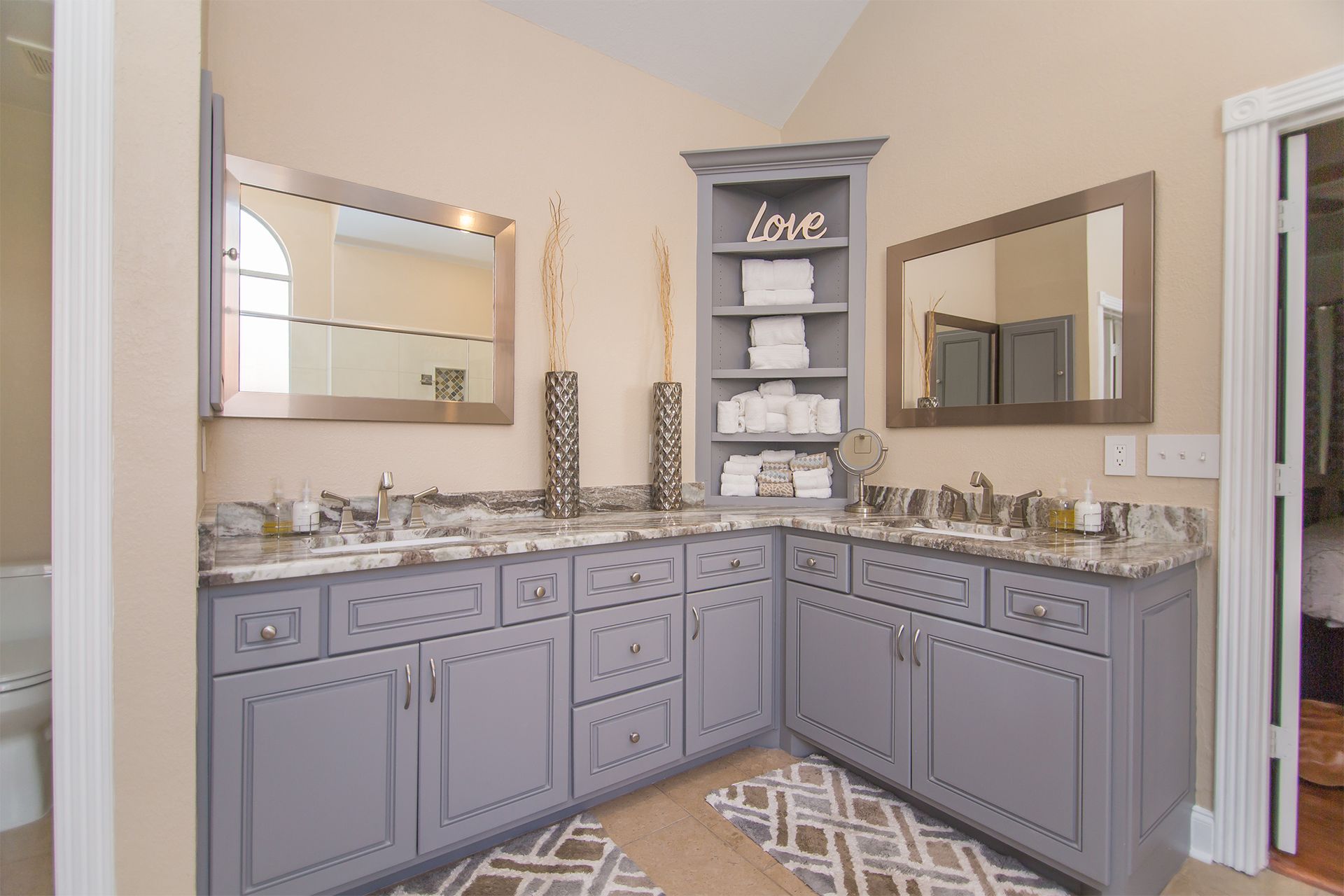 Gray vanity with granite countertops, mirrors, and corner shelving in a beige bathroom. - After