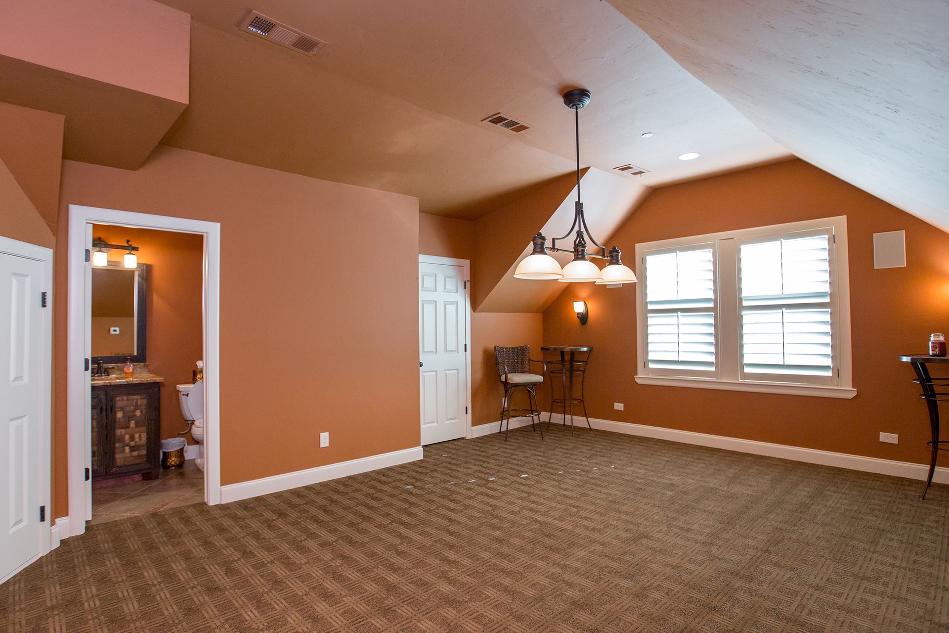 Angled room with orange walls, carpet, and a window. Contains a bathroom door, a white door, and bar stools. - After