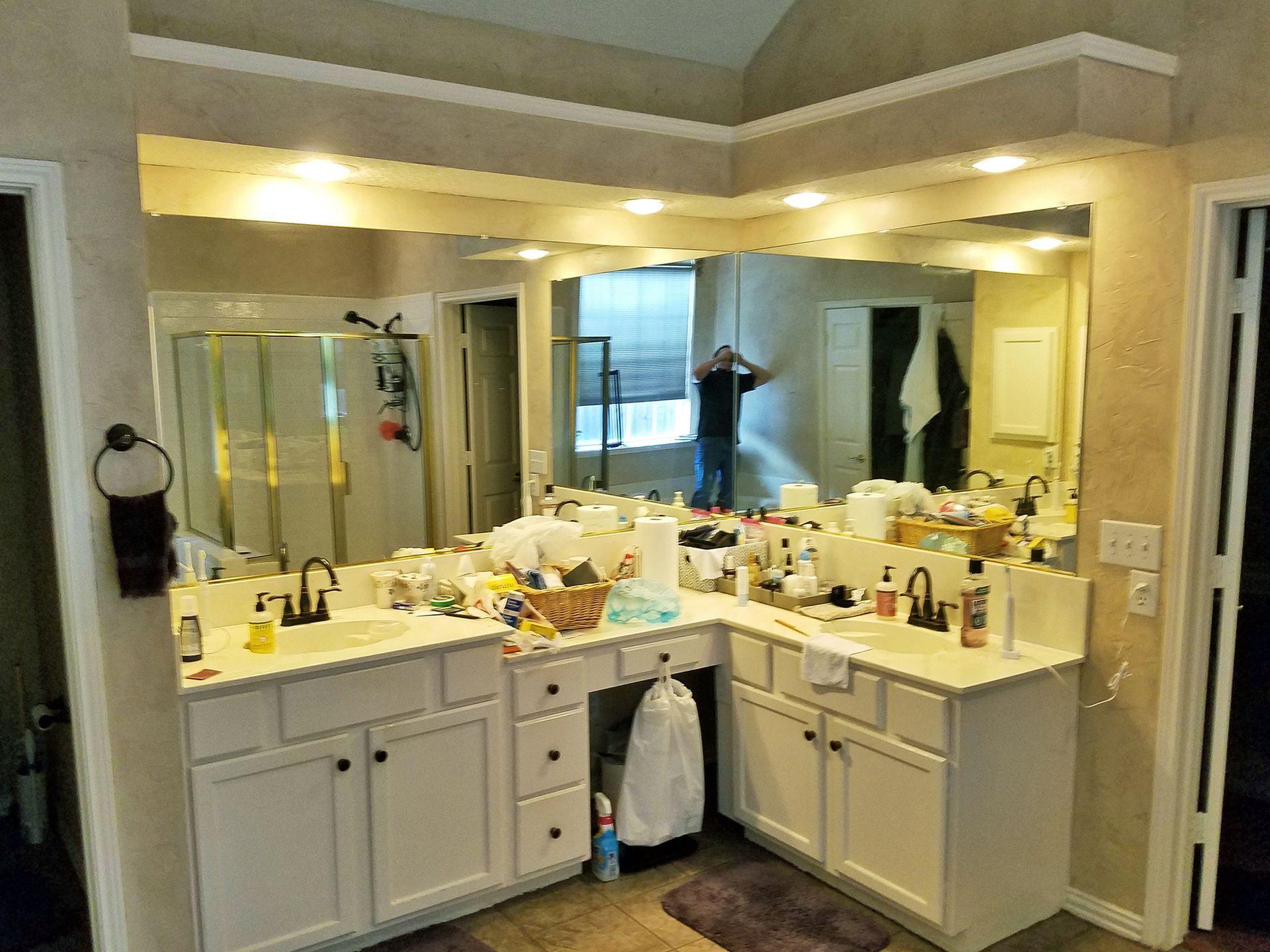 Bathroom with a large mirror, dual sinks, and cluttered countertops; person reflected in the mirror. - Before