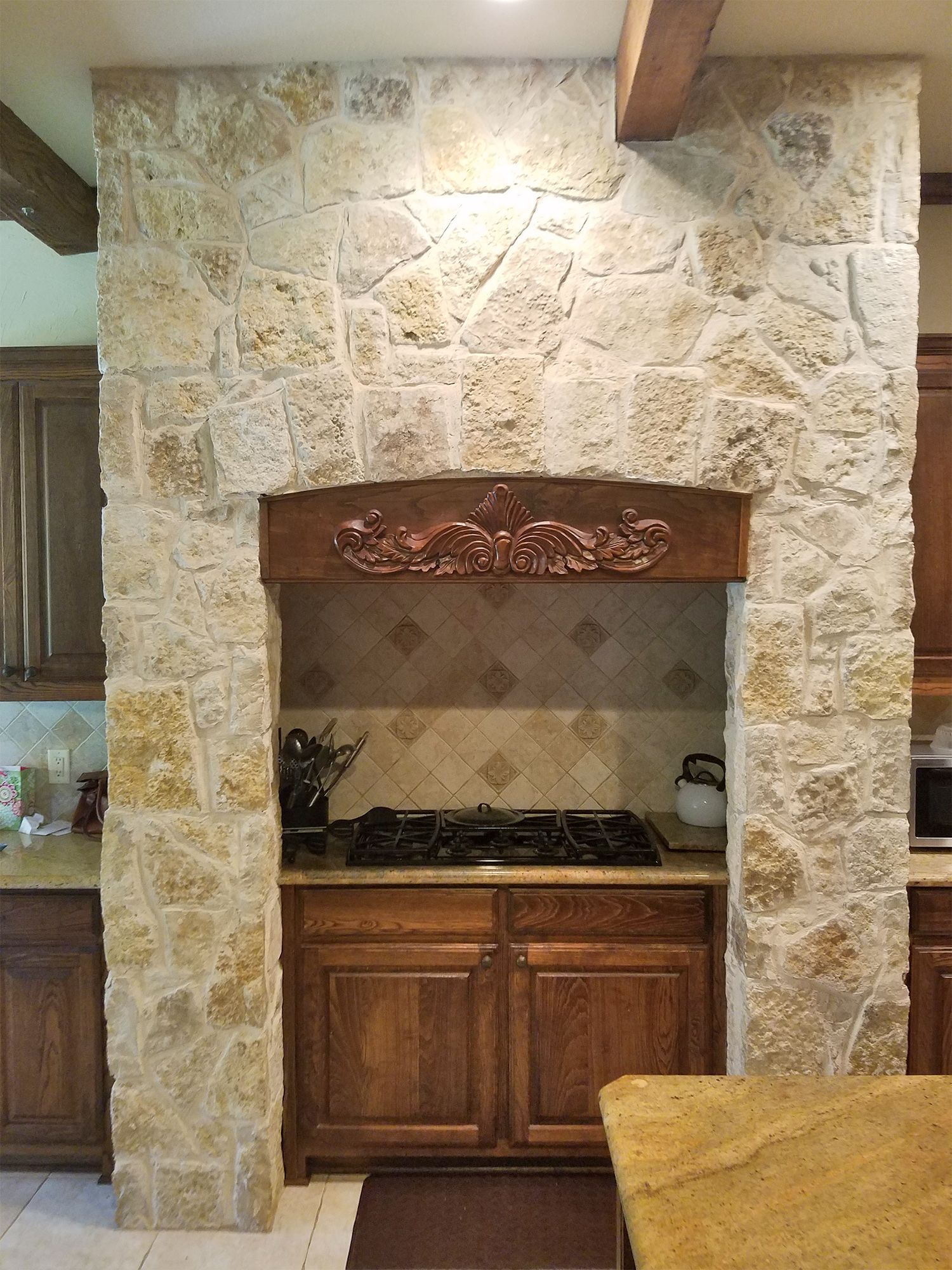 Stone-surrounded cooktop niche with wooden cabinets and brown trim. Beige stone contrasts with the dark wood tones. - Before