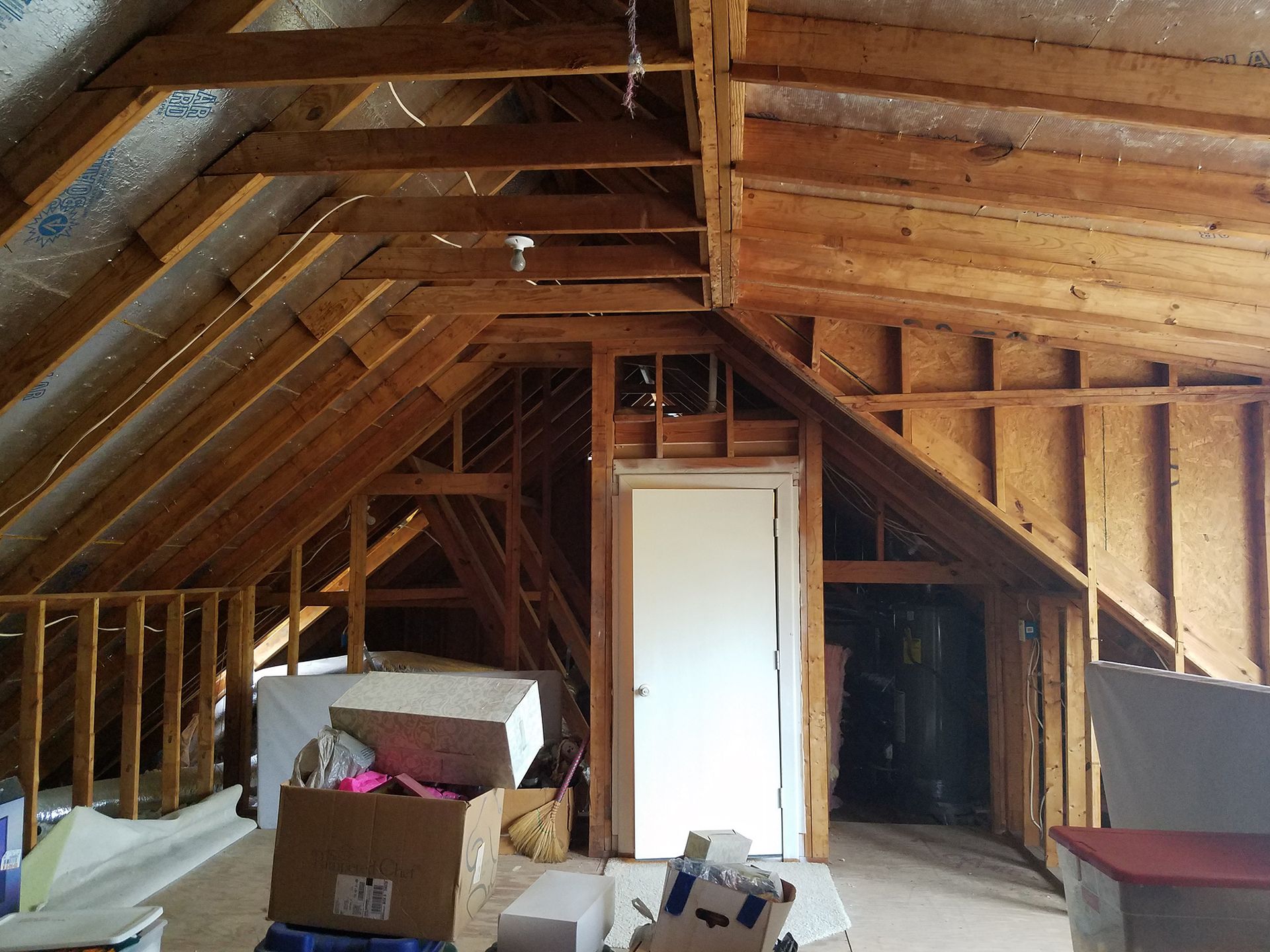 Attic interior with wooden beams, a white door, boxes, and insulation. - Before