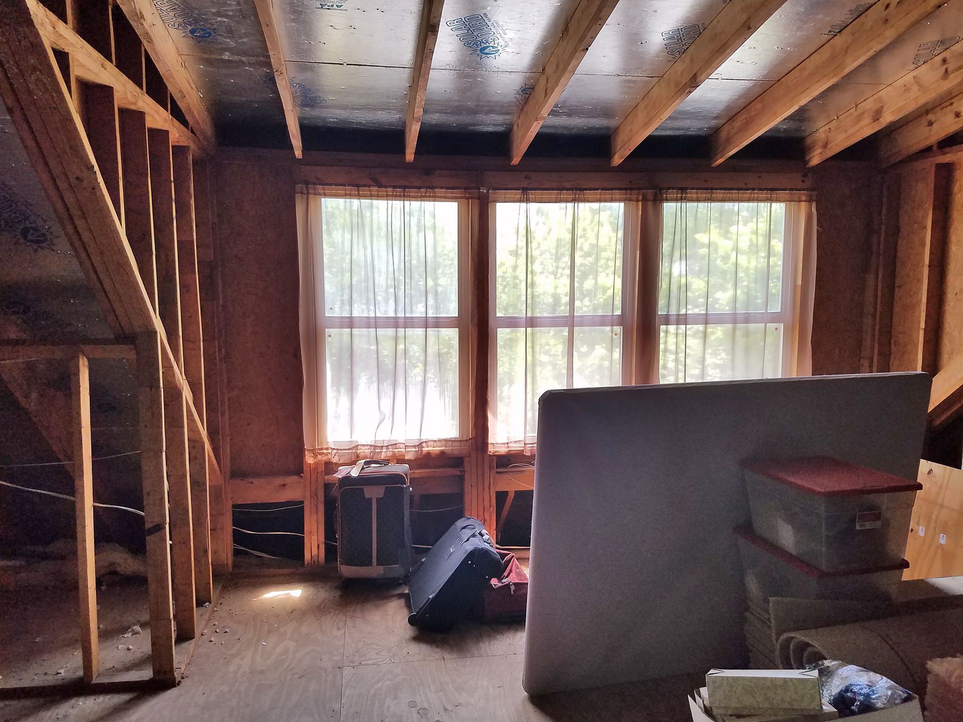 Attic room with sloped walls, windows with sheer curtains, and storage including a mattress, boxes, and a space heater. - Before