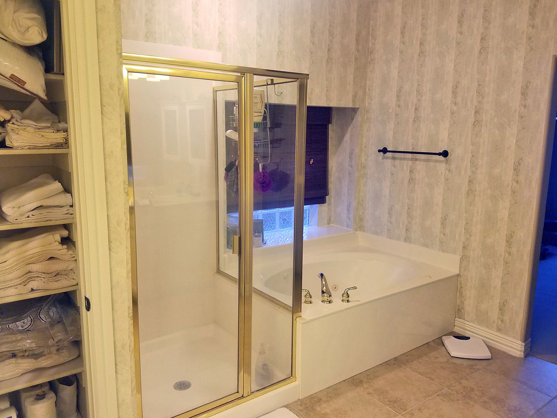 Bathroom with a shower and tub, gold trim, neutral wallpaper and storage shelves. - Before
