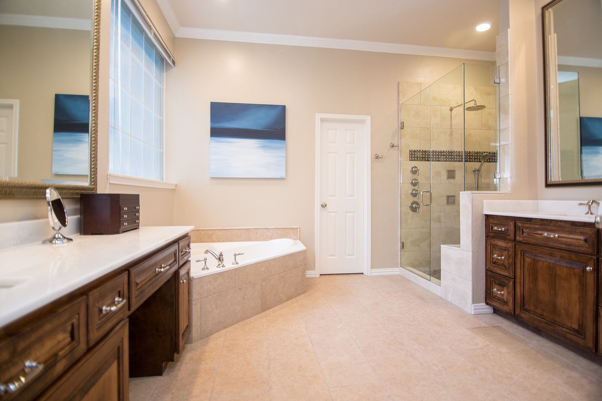 Spacious, beige-toned bathroom with dark wood vanity, glass shower, and a tub.  Modern decor with artwork.