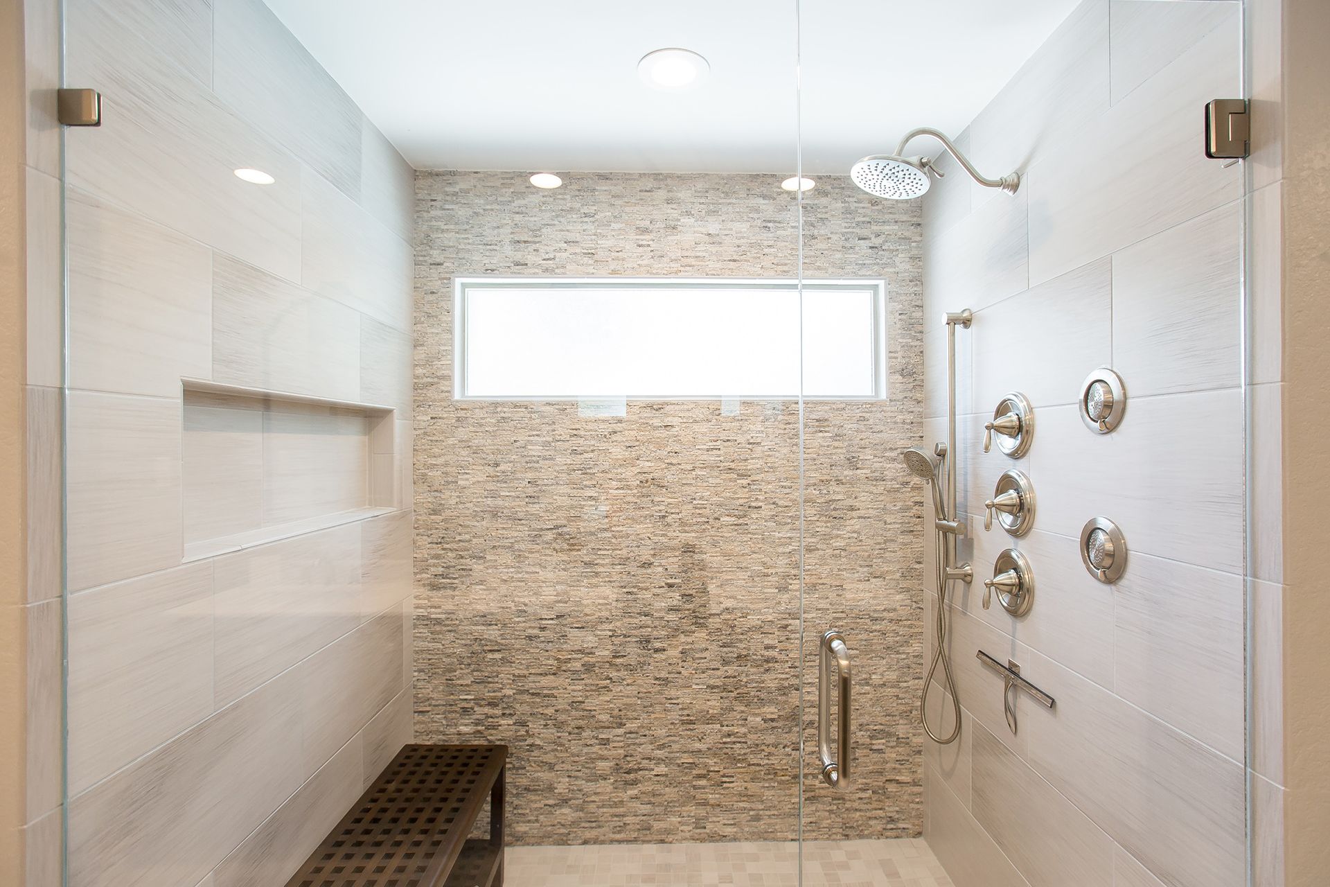 Modern shower with glass doors, textured tile accent wall, bench, and multiple showerheads.