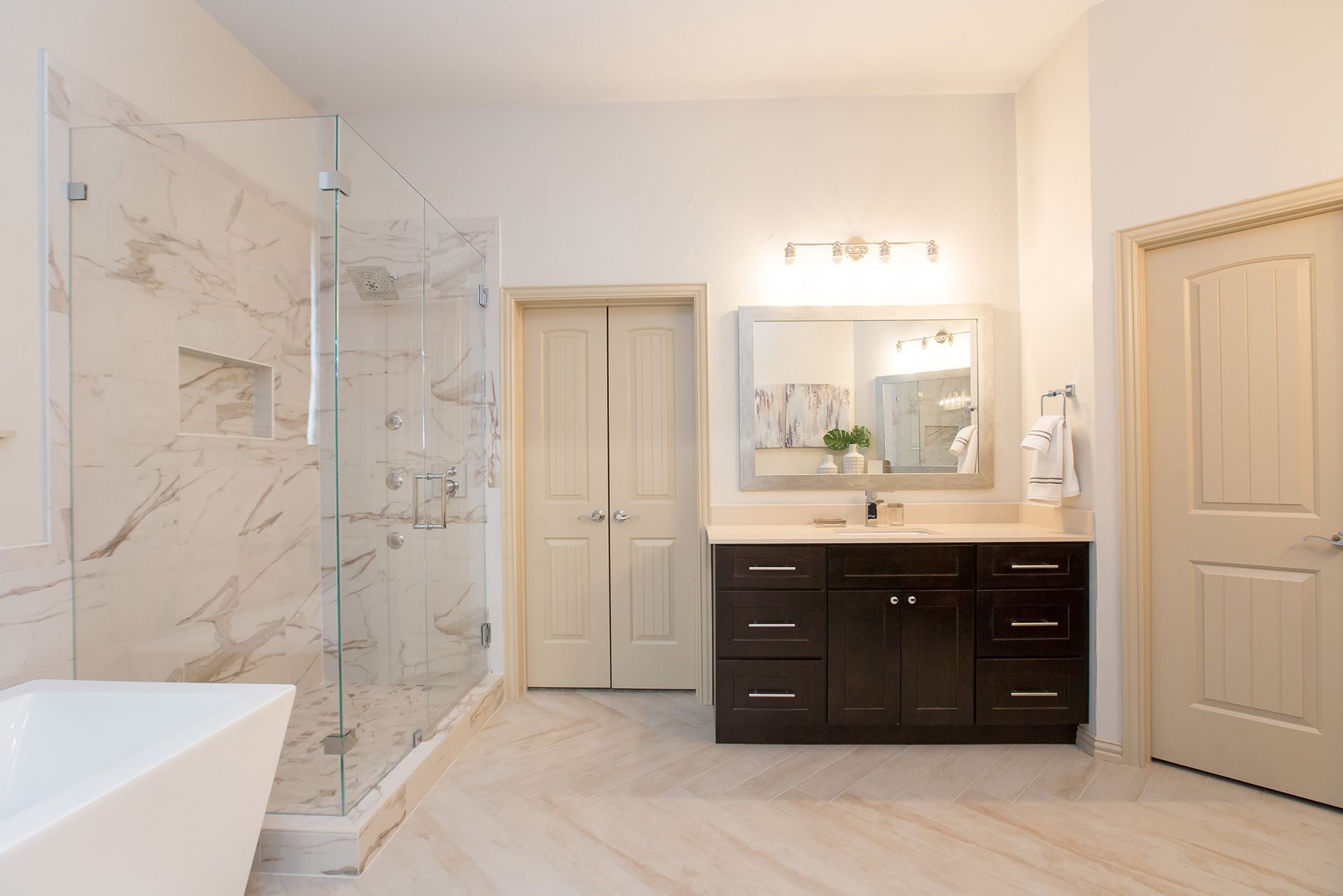 Spacious bathroom with marble shower, dark brown vanity, and two closed doors. White walls and light tile floor.