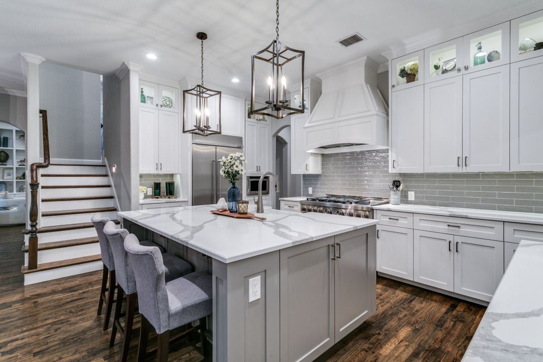 Modern white and gray kitchen with island, pendant lights, and staircase in the background.