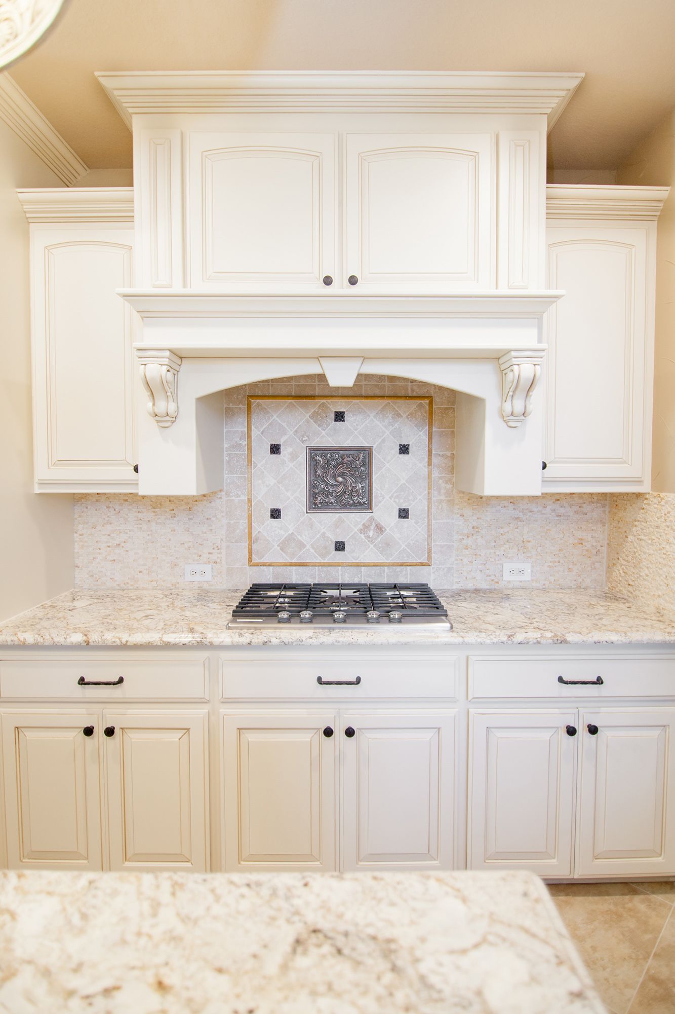 White kitchen cabinets and range hood over a gas stovetop, beige backsplash and countertops.