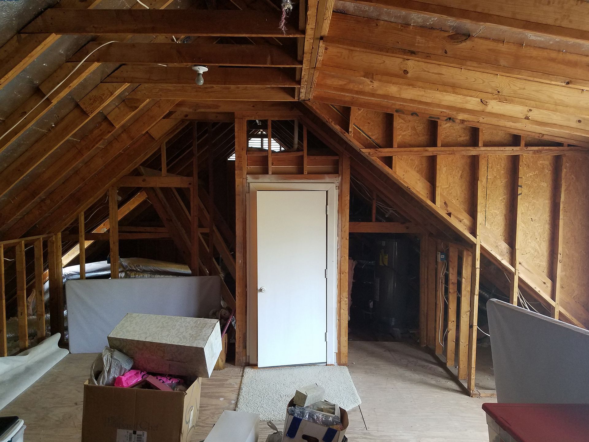 Attic interior with exposed wooden beams and framing. A white door and storage spaces are visible.