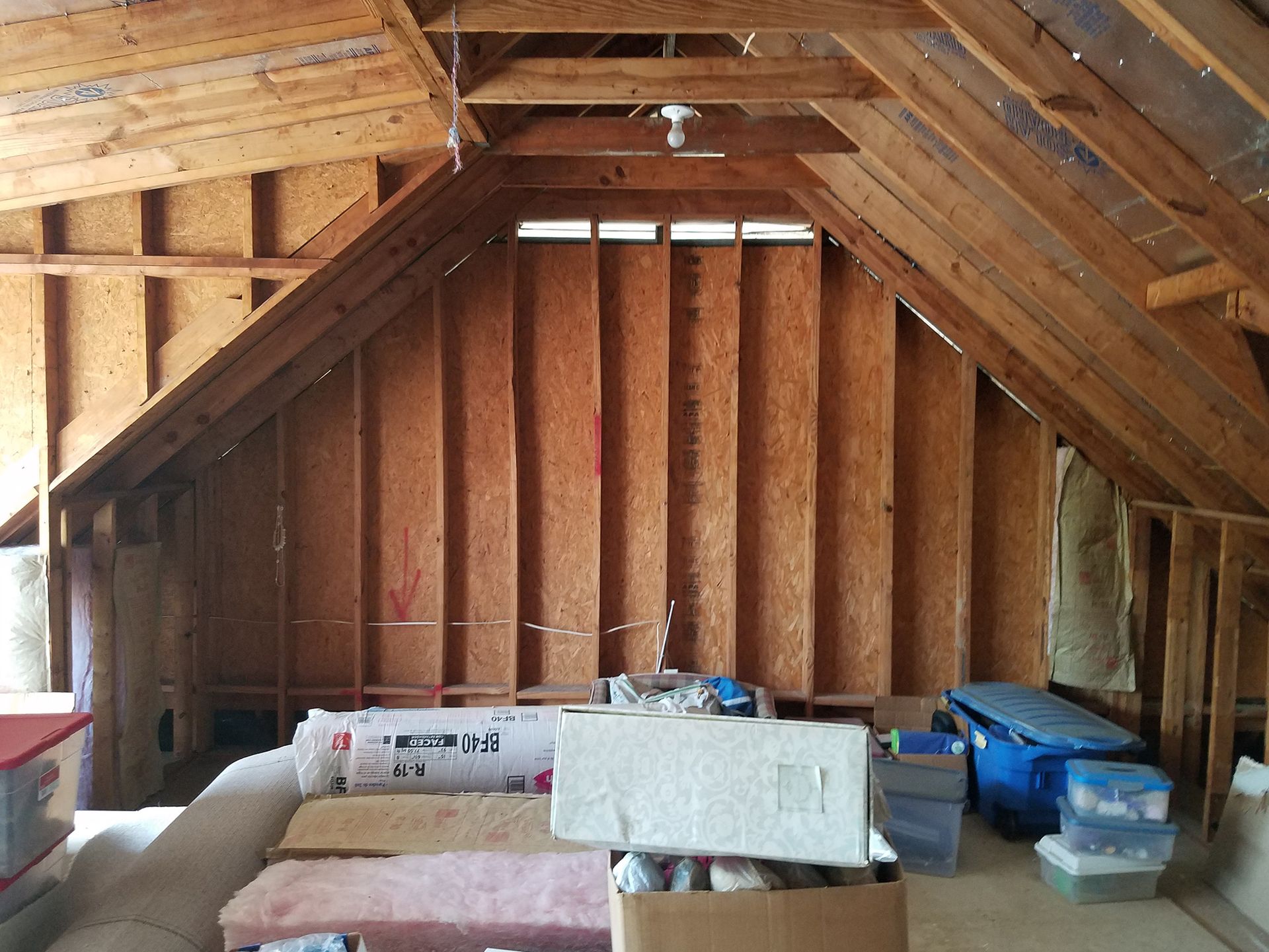 Unfinished attic with exposed wood framing and insulation; materials and boxes clutter the floor.