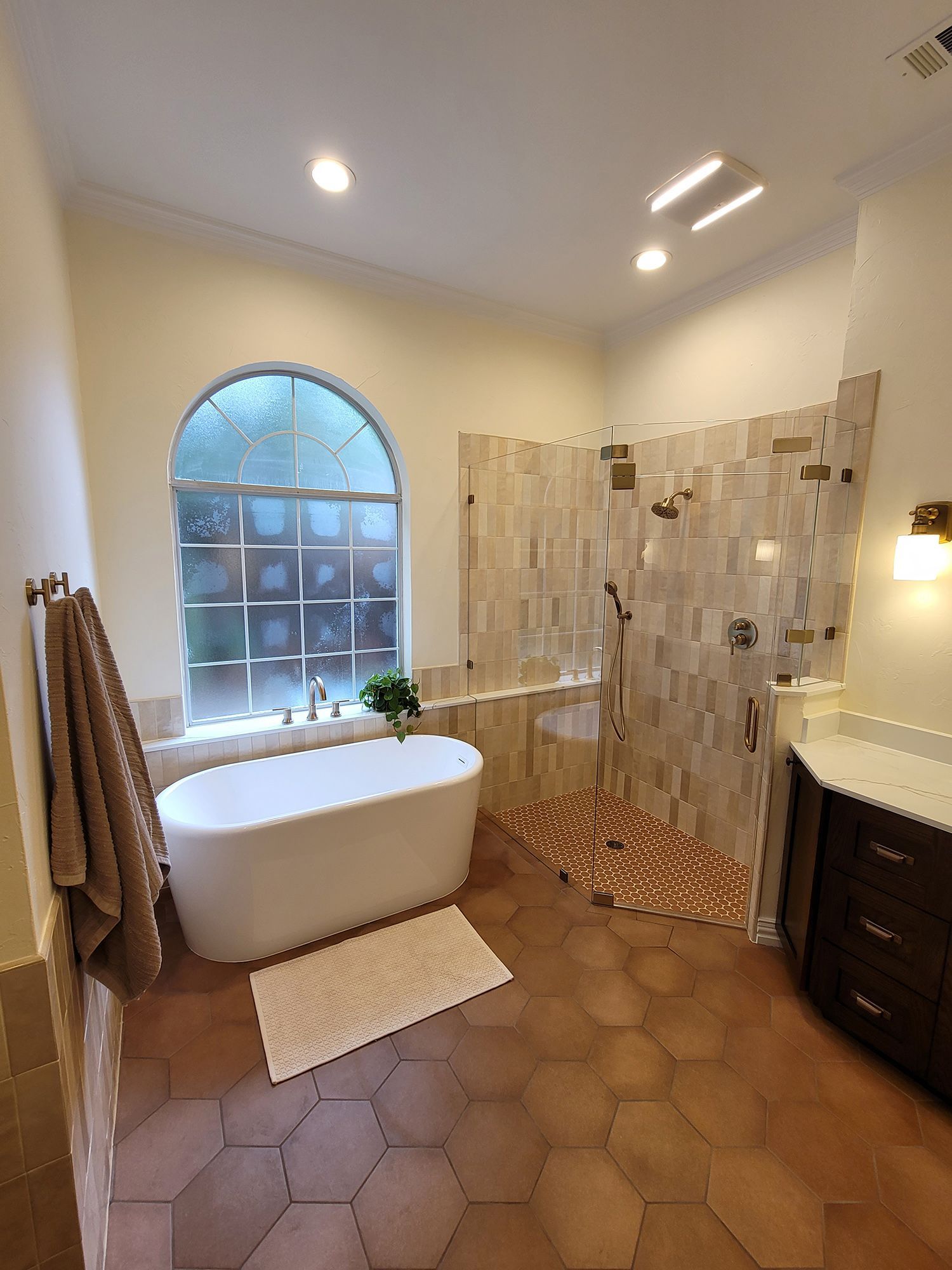 Elegant bathroom: freestanding tub, arched window with glass block, walk-in shower, brown hexagon tiles, and dark vanity.