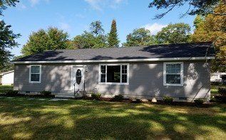 A house with a gray siding and a black roof is sitting on top of a lush green field.