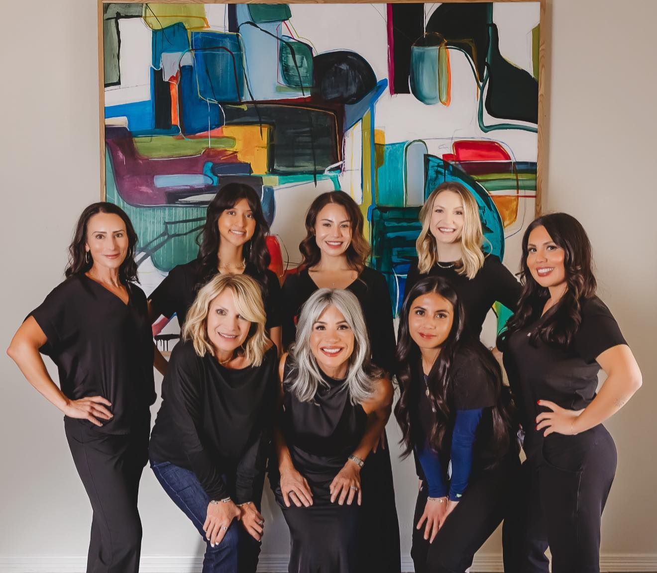 Group of women smiling, posing in front of colorful abstract art. They wear dark clothing, posing indoors.