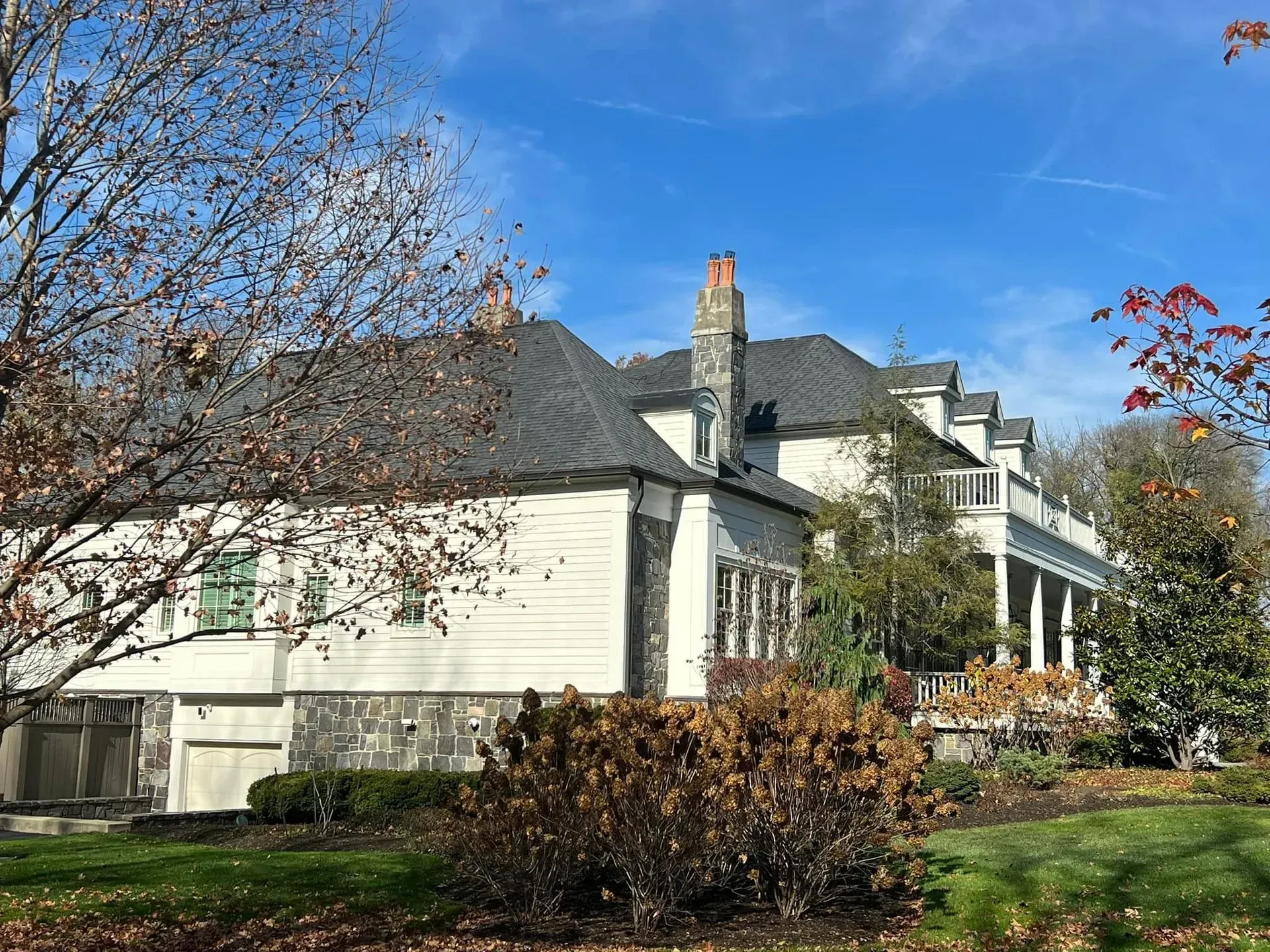 A large white house with a gray roof is surrounded by trees and bushes on a sunny day.