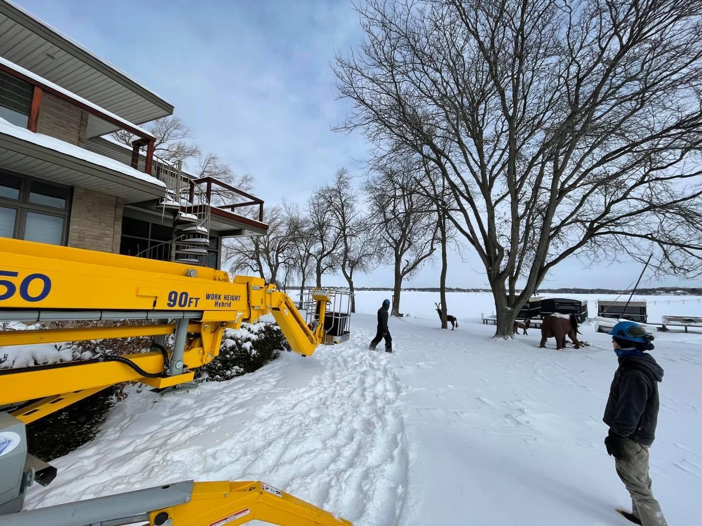 Snowy scene: Yellow lift near a building, people walking on snow, bare trees, lake in background.