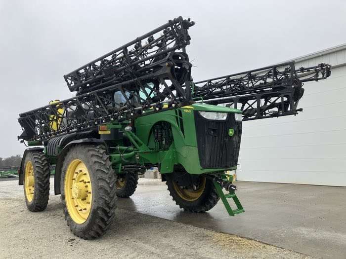 Green agricultural sprayer with large black booms and yellow wheels, parked on wet pavement outdoors