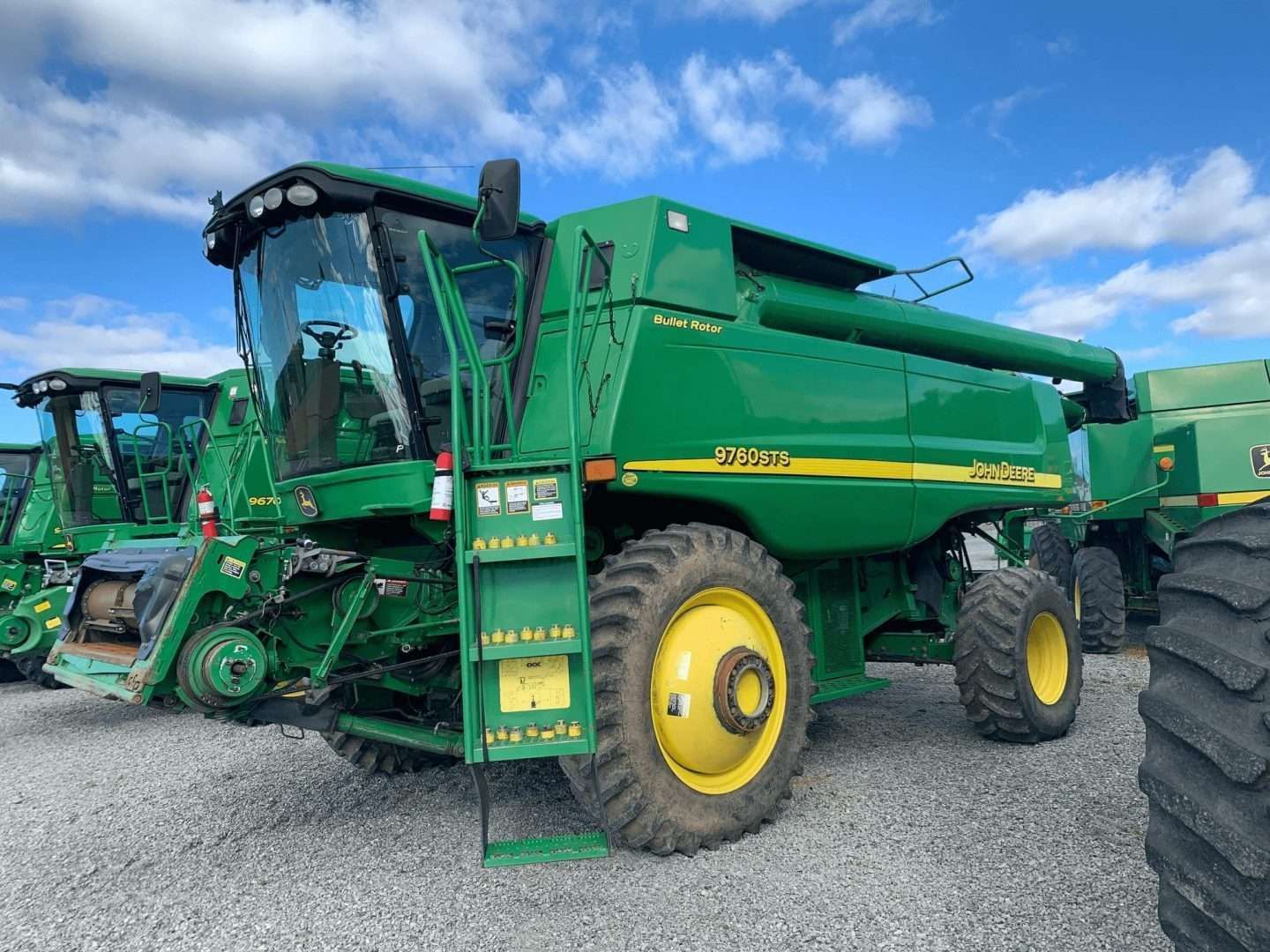 Green combine harvester parked on gravel under a blue sky, with another machine beside it.