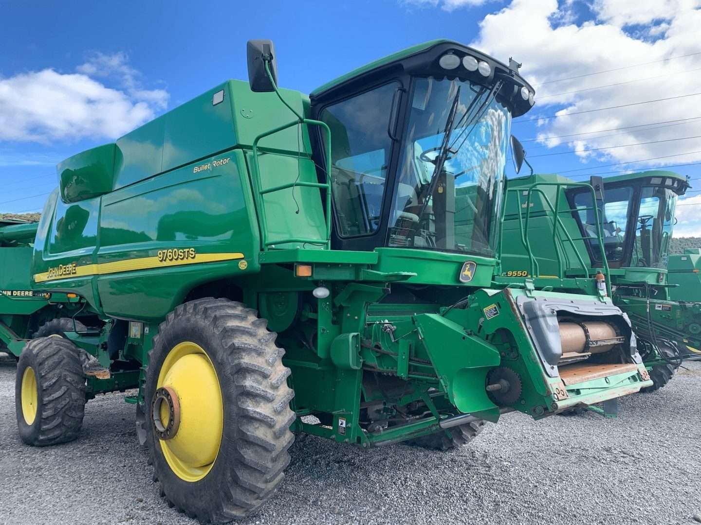 Green combine harvester on a gravel lot under a blue sky, with another machine in the background.