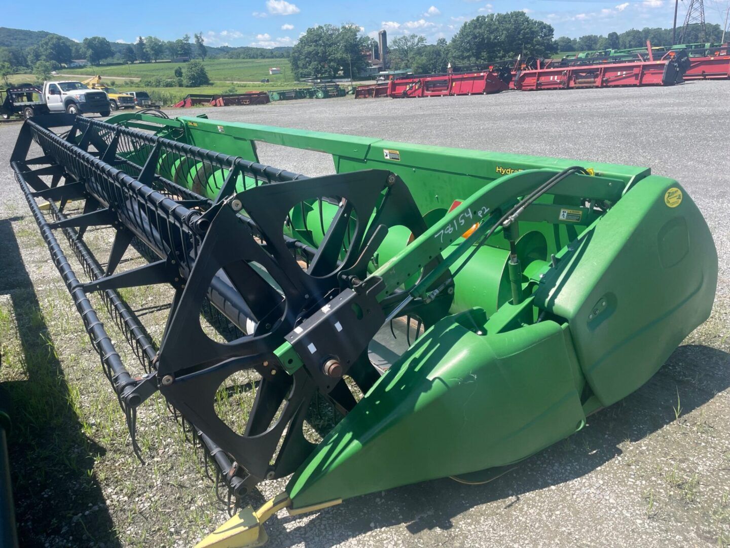 Green agricultural header attachment parked outdoors on gravel, with black reel and red farm equipment in the background