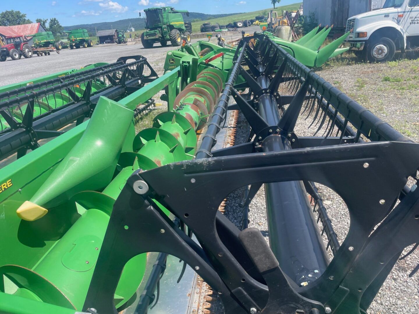 Green farm combine headers lined up outdoors on gravel, with trucks and fields in the background.