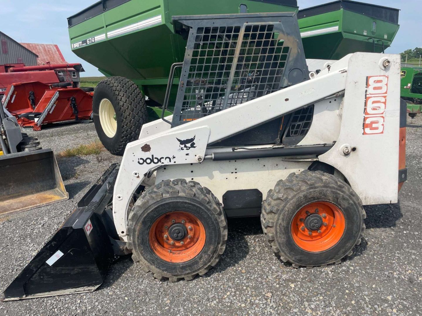 Bobcat skid steer with black bucket and orange wheels parked beside green farm equipment.