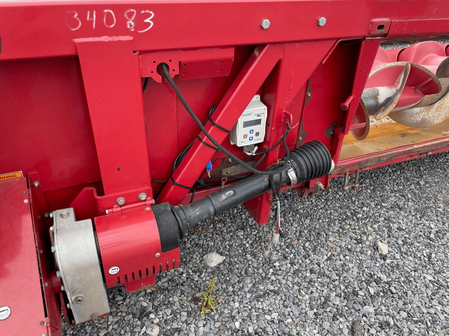 Red farm machine hitch with hydraulic cylinder and black hoses on gravel.