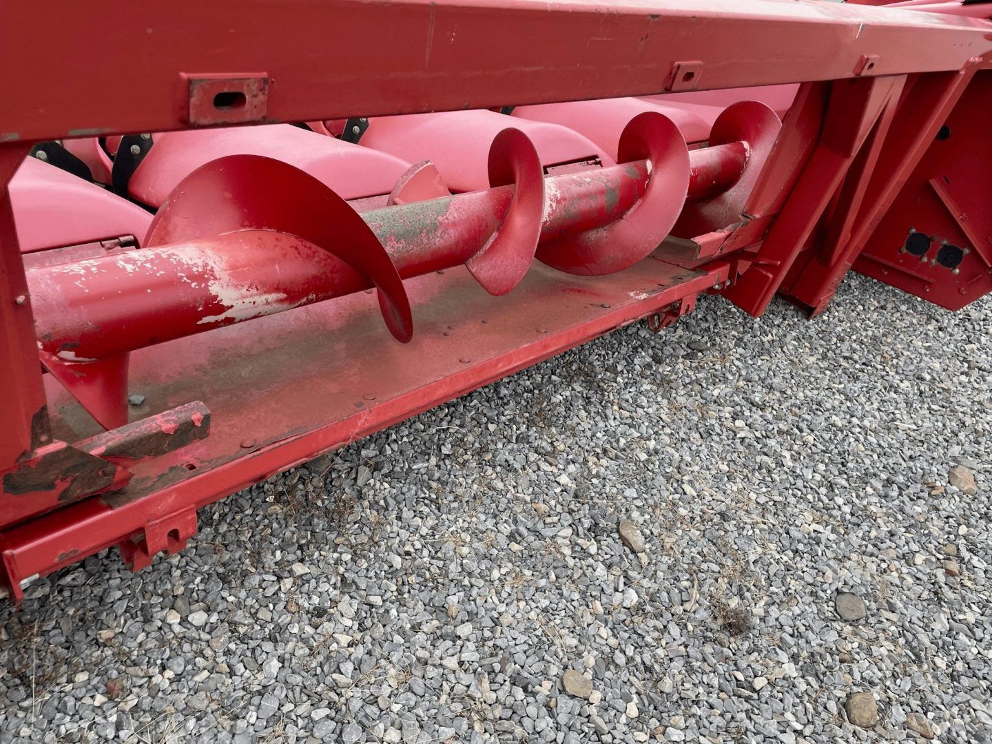 Red farm machine with a large metal auger on gravel ground