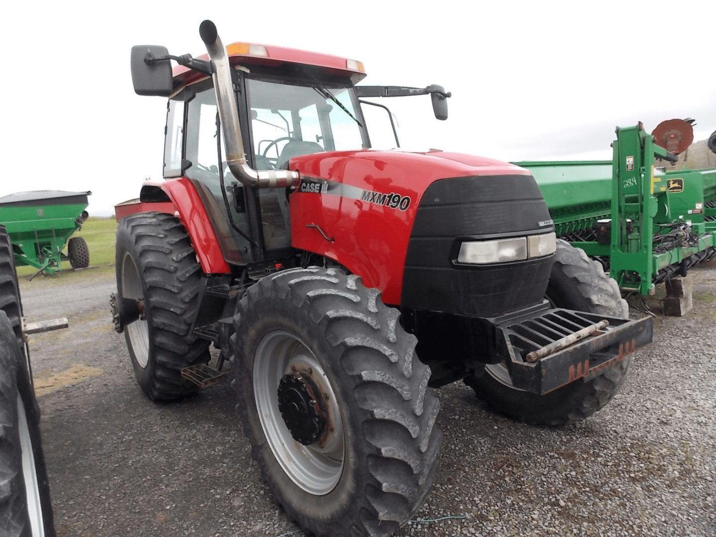 Red tractor parked on gravel lot with green farm equipment in the background.