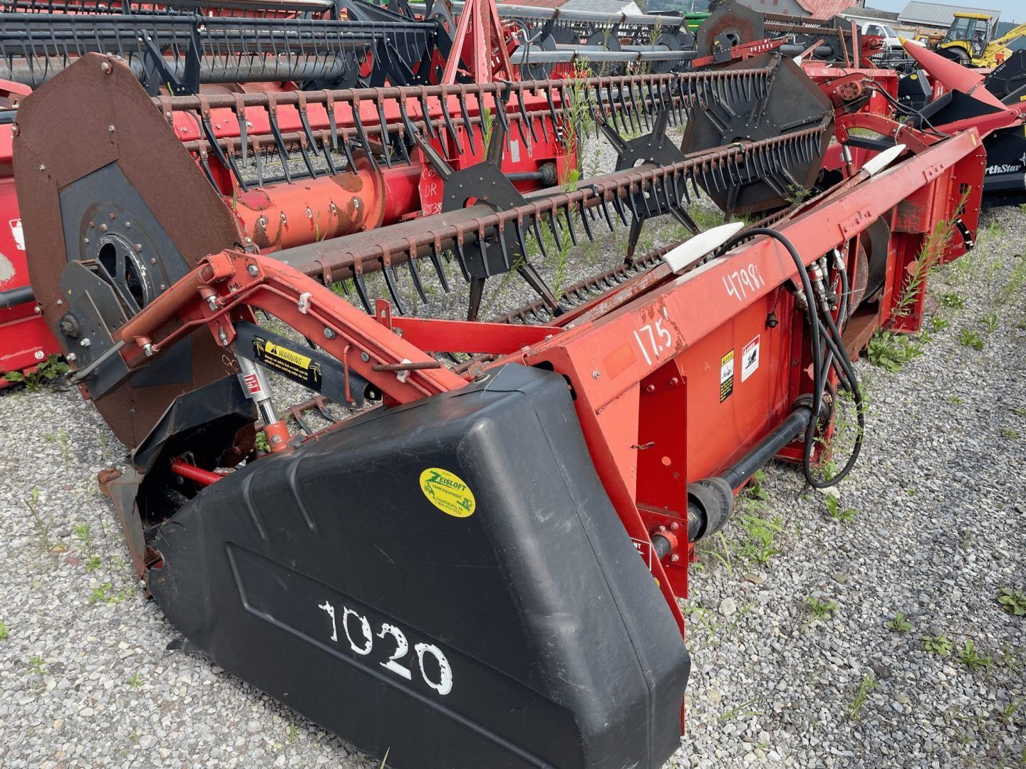 Red farm combine header with black end guard, sitting on gravel lot with other equipment behind it
