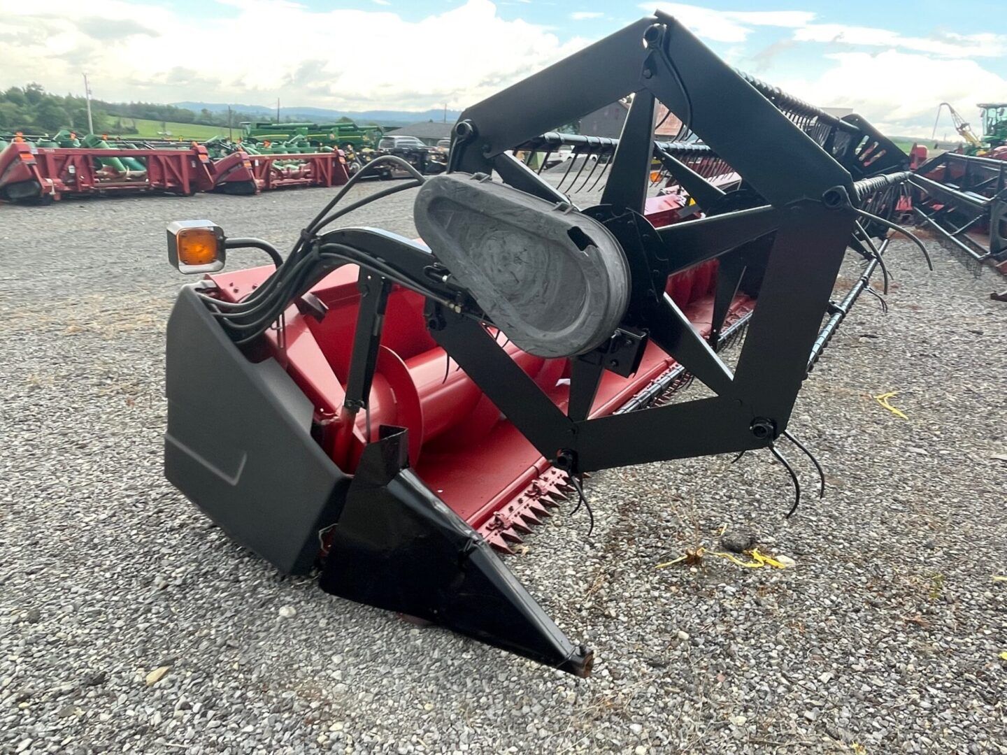 Red agricultural machine attachment with black frame and red body on gravel lot, rows of equipment behind it