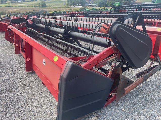 Red farm machinery attachment with black conveyor belt, parked on gravel in a field