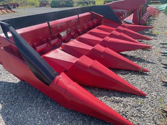 Red corn harvester header with pointed metal teeth on gravel outdoors