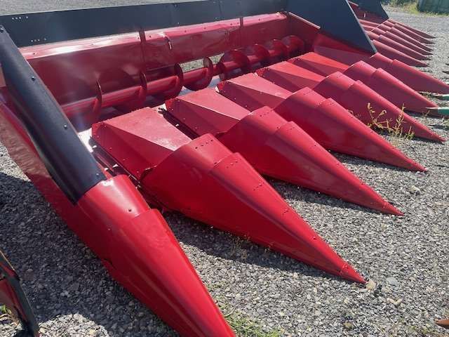 Close-up of red corn header harvesting attachment with pointed metal prongs on gravel.