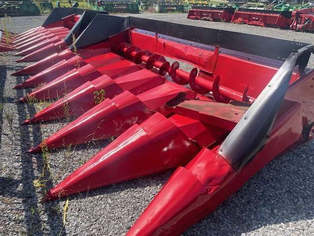 Red farm combine header with rows of pointed corn snouts on gravel lot
