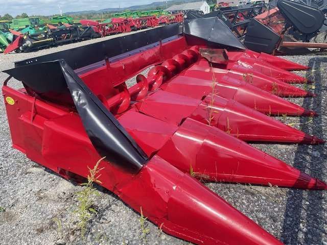 Red agricultural header with multiple pointed red dividers and black guards, parked outdoors on gravel.