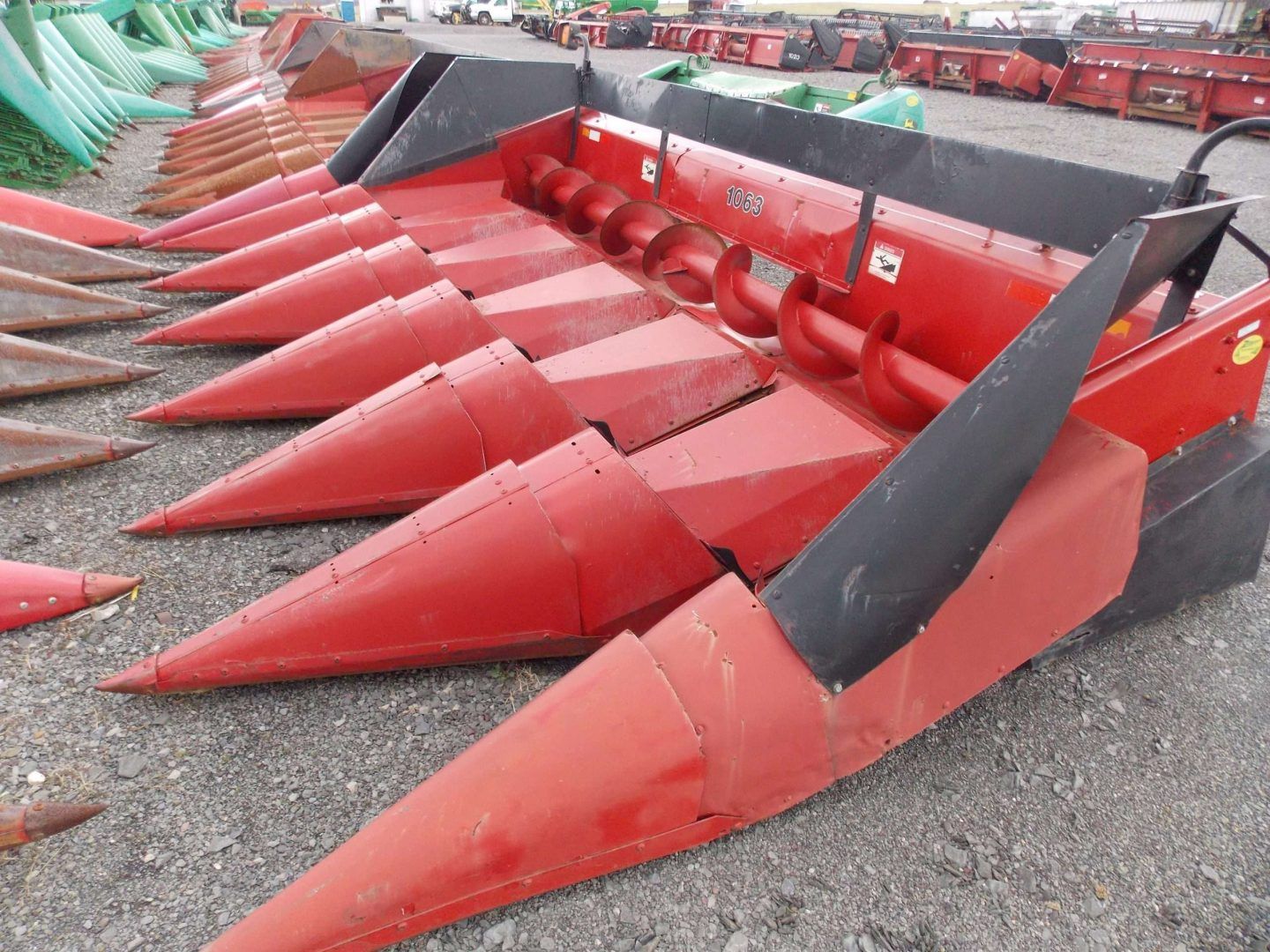 Red corn harvester header with multiple pointed metal rows on a gravel lot