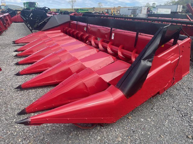Red agricultural corn harvester header with pointed red rows and black guard arms on gravel lot