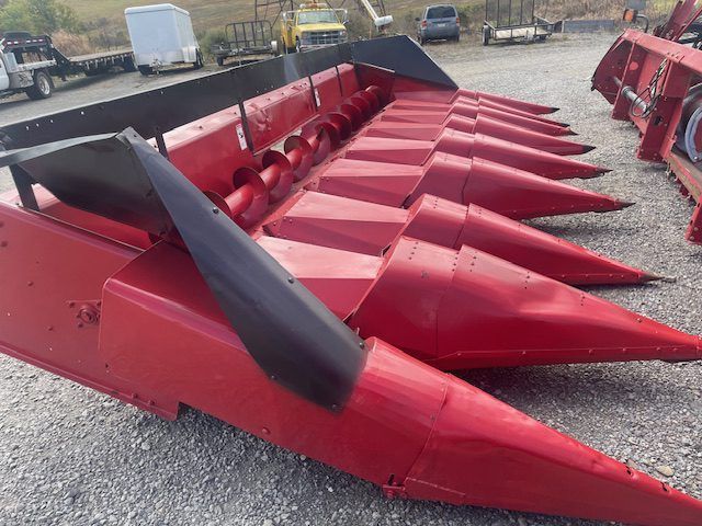 Red farm combine corn header with pointed metal crop dividers on gravel lot