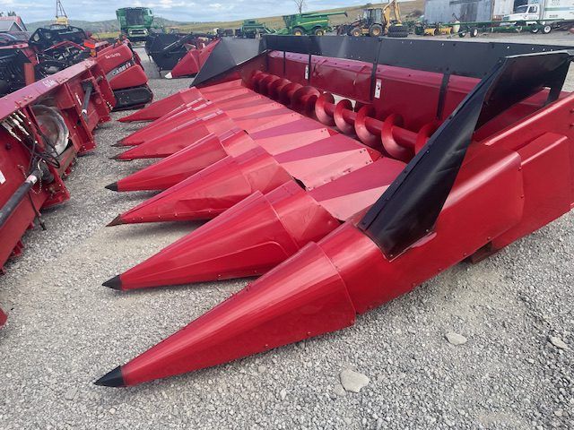 Red corn header with pointed red snouts and black toppers lined up on gravel outdoors