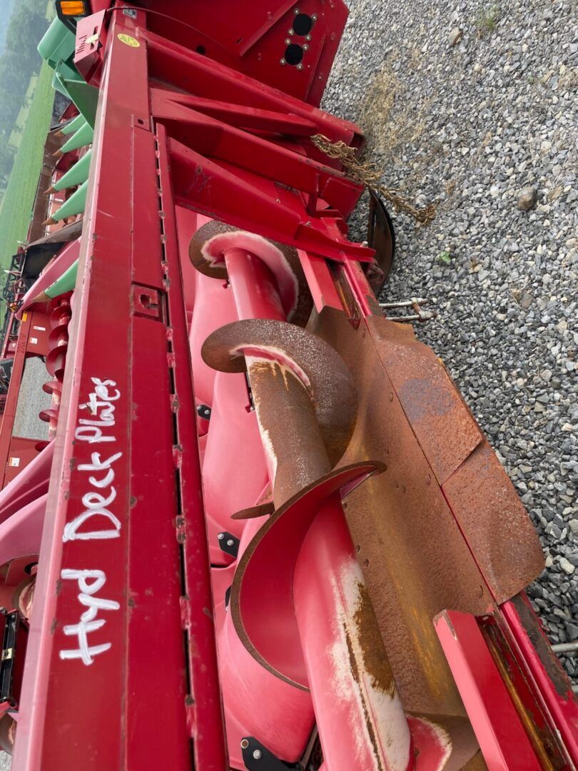 Red farm equipment with red metal blades and white writing on the side, parked on gravel.