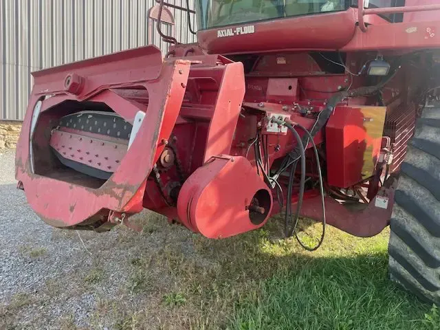 Red farm machinery with exposed internal parts and a large wheel, parked on gravel outdoors.