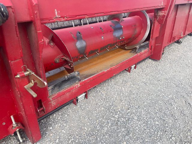 Red industrial machine with a cylindrical drum over a wooden plank on gravel.