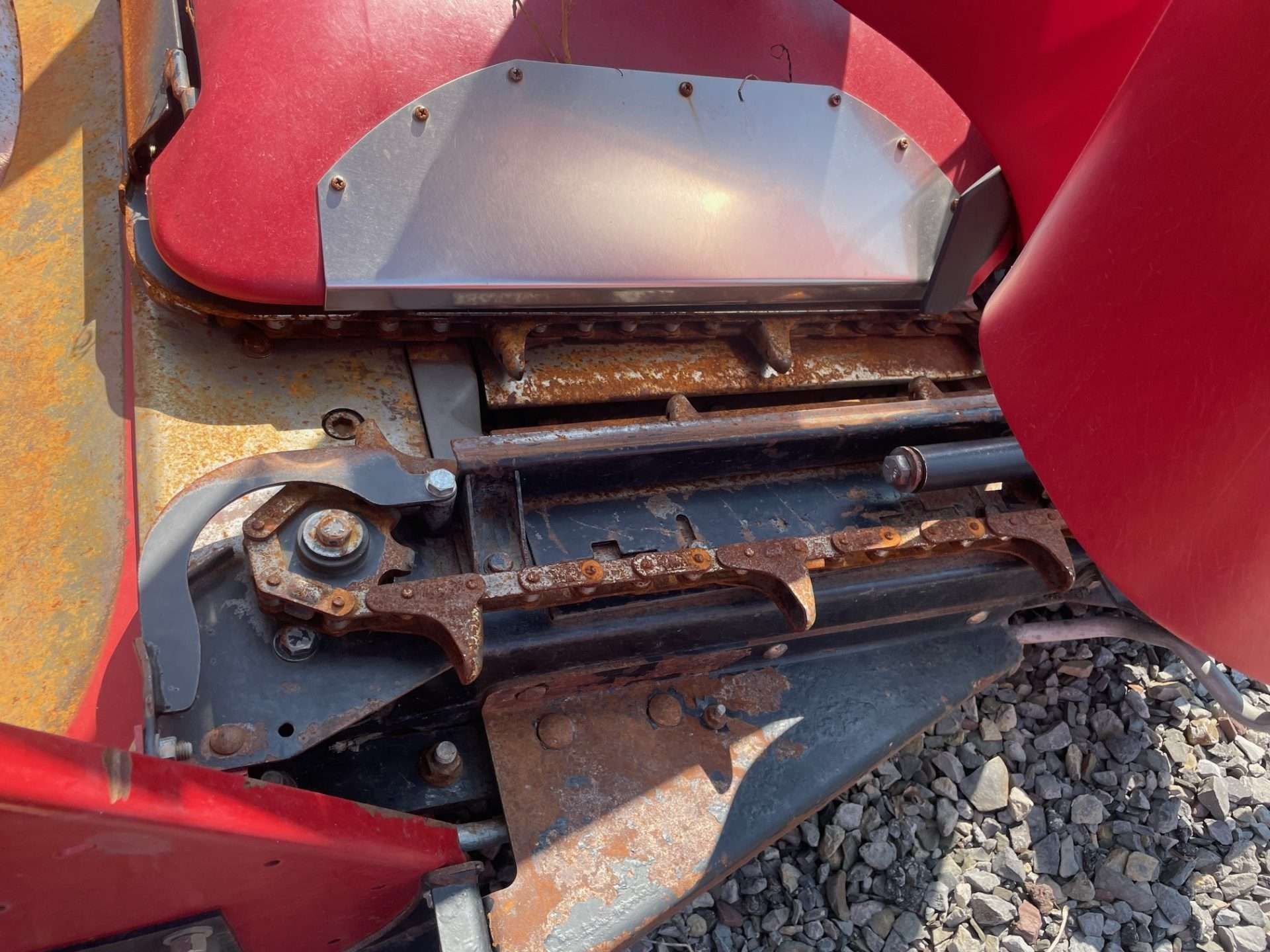 Close-up of a red agricultural machine’s worn metal hinge and track over gravel.