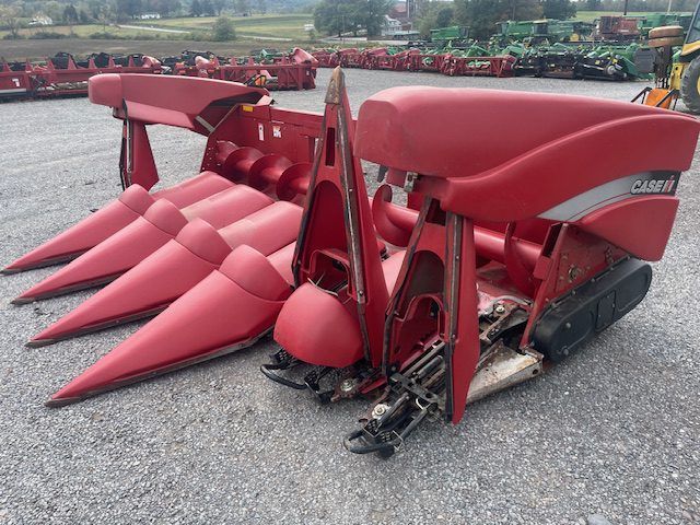 Red farm combine header with multiple pointed corn rows, parked on gravel in a field lot